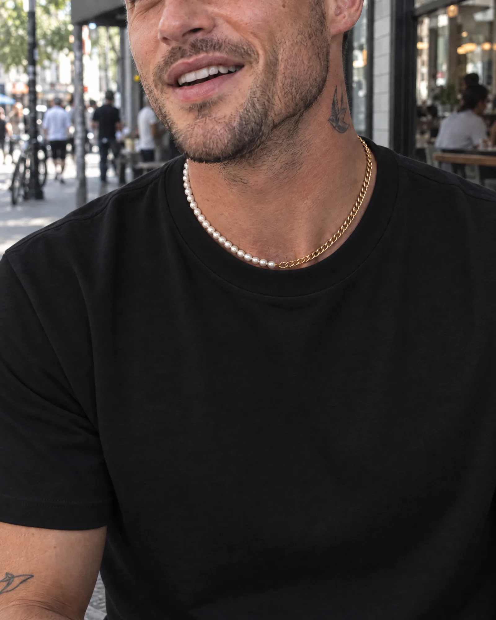 A man in a plain black T-shirt, a small tattoo on his neck, wears the pearl and gold necklace outside a café in daylight.