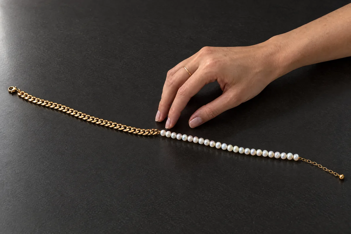 A woman's hand reaches for the pearl and gold necklace lying on black stone.