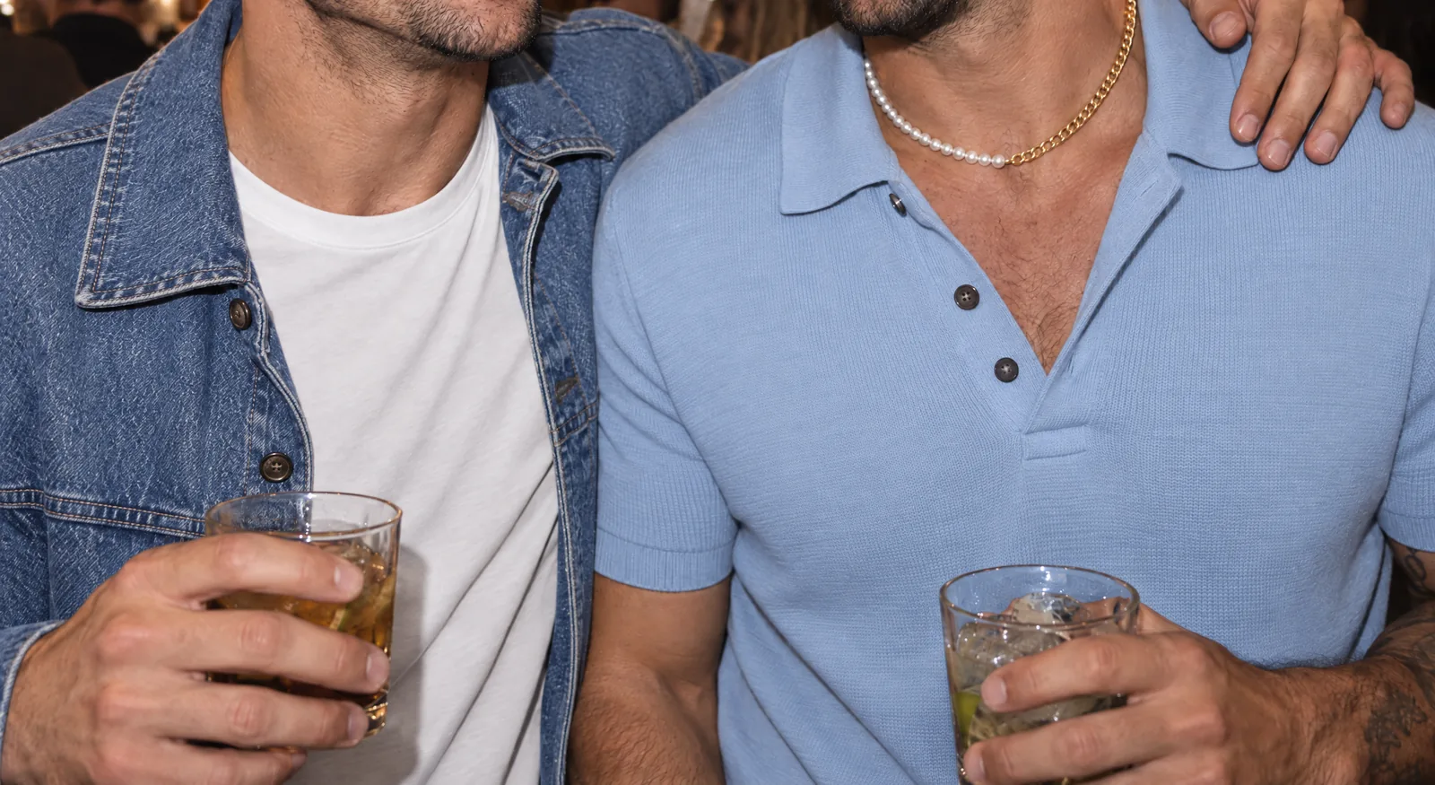 Two friends at a busy bar at night, seen from the chin down, one in a denim jacket with an arm round the other's shoulder; the one in the blue knit polo wears the pearl and gold necklace.